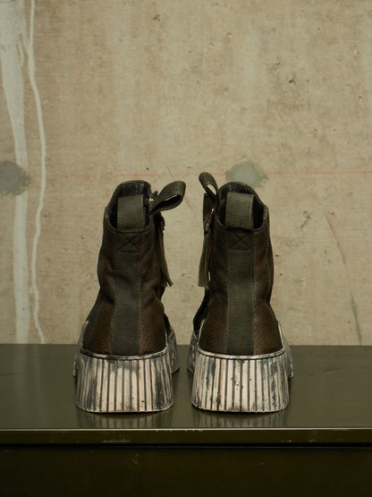 A pair of dark brown leather ankle boots with distressed silver platform soles, shown from the back on a dark table against a textured concrete wall.