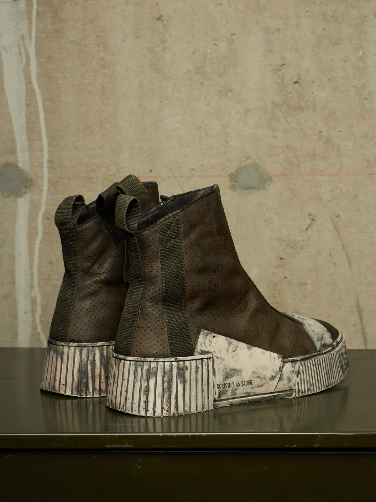 A pair of high-top boots with dark brown textured leather uppers and chunky, ribbed platform soles in a distressed off-white and gray pattern, displayed on a dark surface against a textured concrete wall.