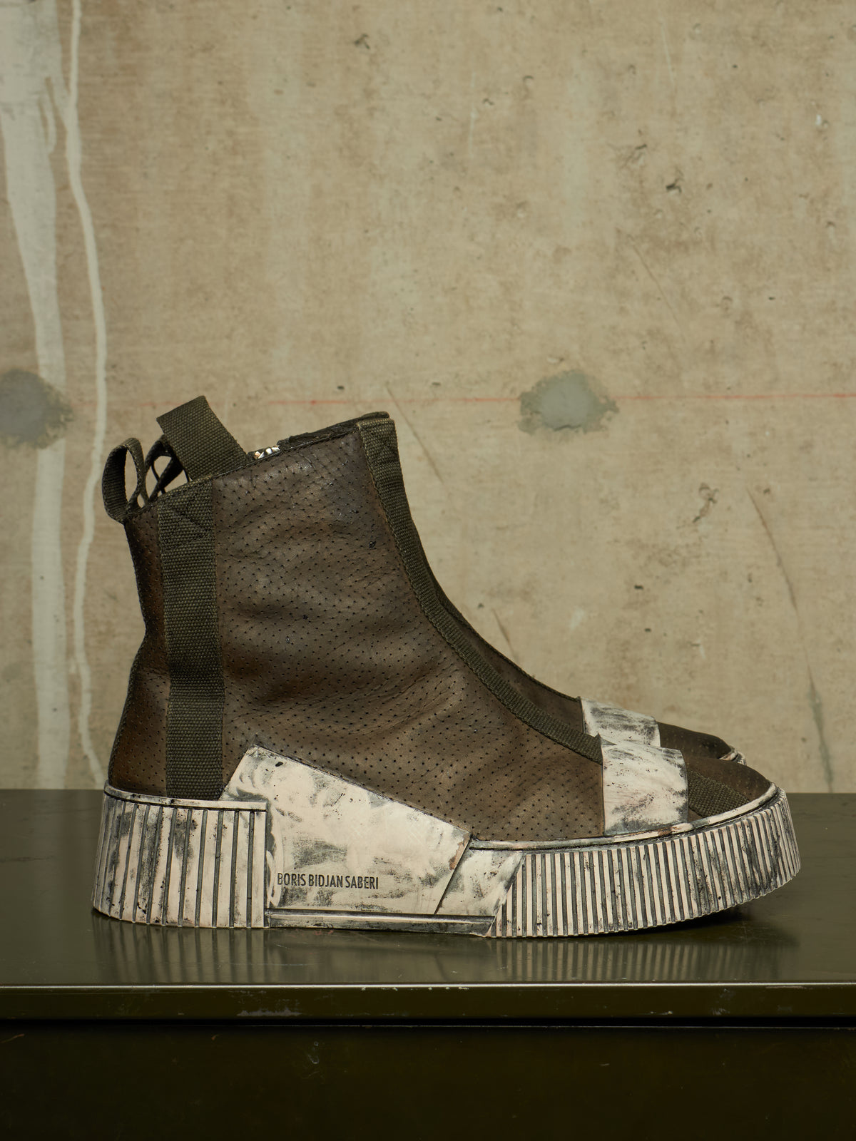 A single high-top sneaker shown in profile on a dark surface against a textured concrete wall. The shoe has a dark brown perforated leather upper with a thick platform sole featuring distressed silver and white panels.