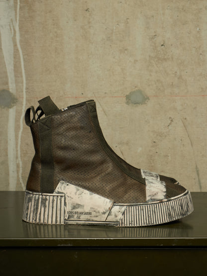 A single high-top sneaker shown in profile on a dark surface against a textured concrete wall. The shoe has a dark brown perforated leather upper with a thick platform sole featuring distressed silver and white panels.