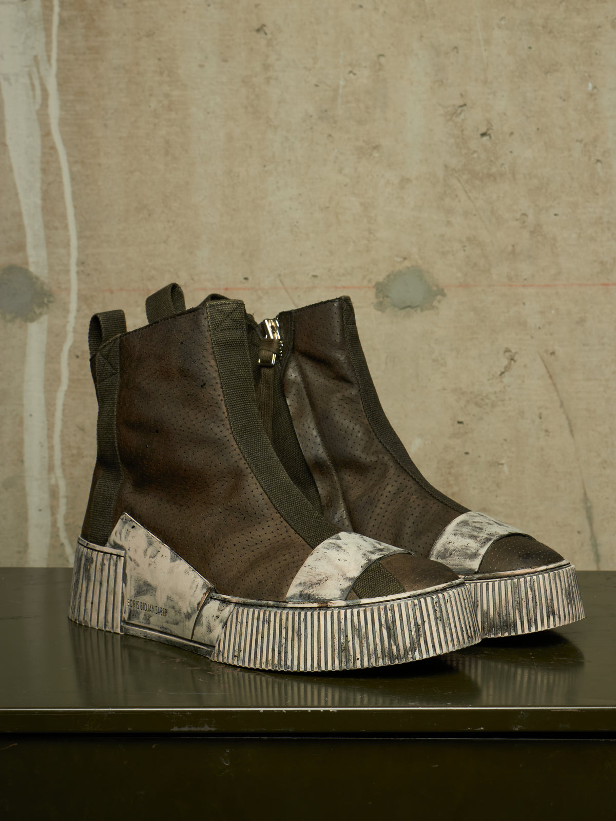A pair of dark brown high-top ankle boots with textured leather panels, side zippers, and chunky striped platform soles, displayed on a dark reflective surface against a textured concrete wall.
