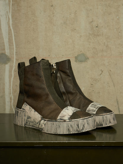 A pair of dark brown high-top ankle boots with textured leather panels, side zippers, and chunky striped platform soles, displayed on a dark reflective surface against a textured concrete wall.