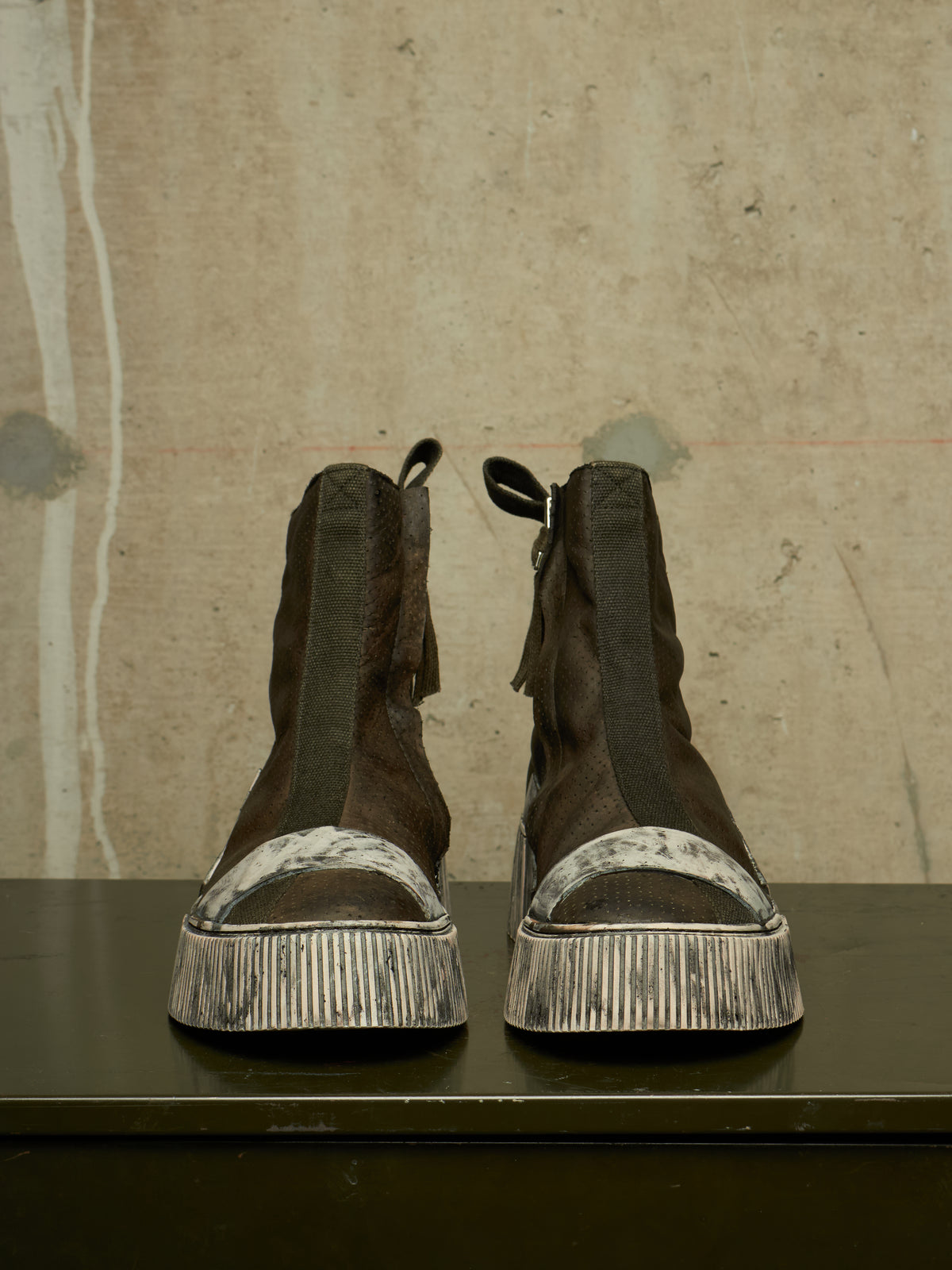 A pair of dark brown suede ankle boots with distressed silver platform soles, displayed on a dark surface against a textured concrete wall.