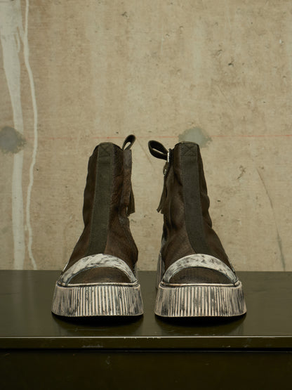 A pair of dark brown suede ankle boots with distressed silver platform soles, displayed on a dark surface against a textured concrete wall.