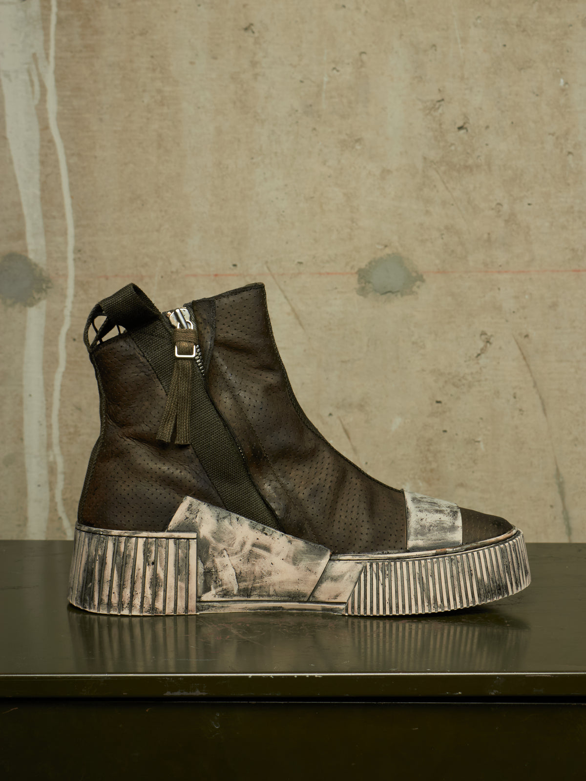 A single dark brown textured ankle boot with a side zipper, tassel detail, and a heavily distressed, painted-outlook sole, displayed on a dark reflective surface against a textured concrete wall.
