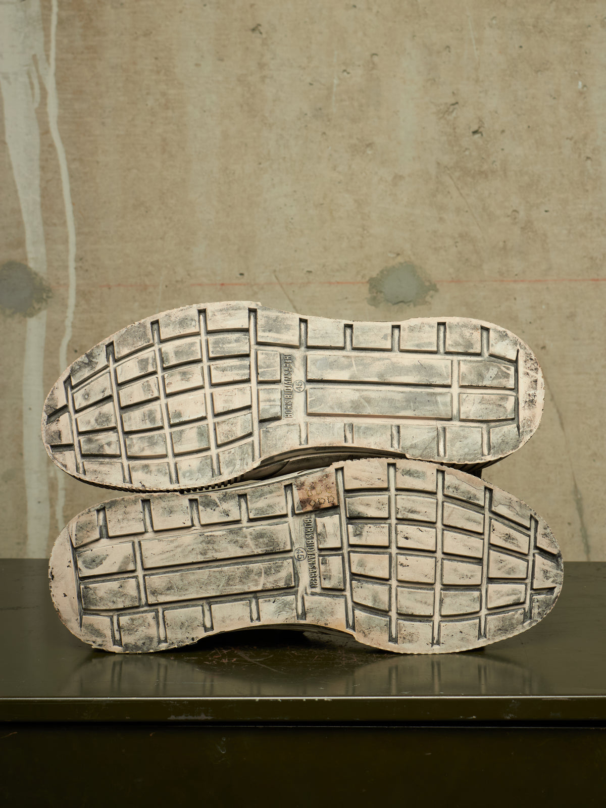 The treaded soles of a pair of shoes stacked vertically on a dark surface against a textured concrete wall.