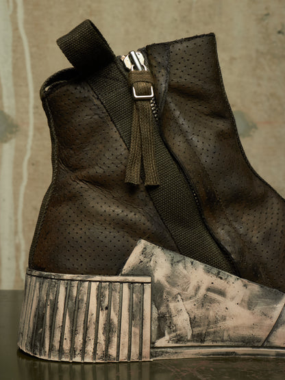 A dark brown perforated leather ankle boot with a chunky, distressed silver and black platform sole, featuring a side zipper with a tassel pull.