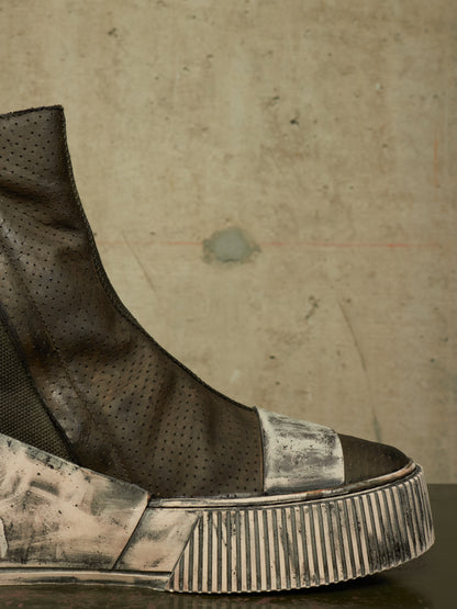 A close-up of a single high-top sneaker with a distressed, weathered finish. The shoe features a dark brown perforated leather upper, a chunky platform sole with a ribbed pattern, and a silver-toned metal toe cap, all showing signs of wear and scuffing.