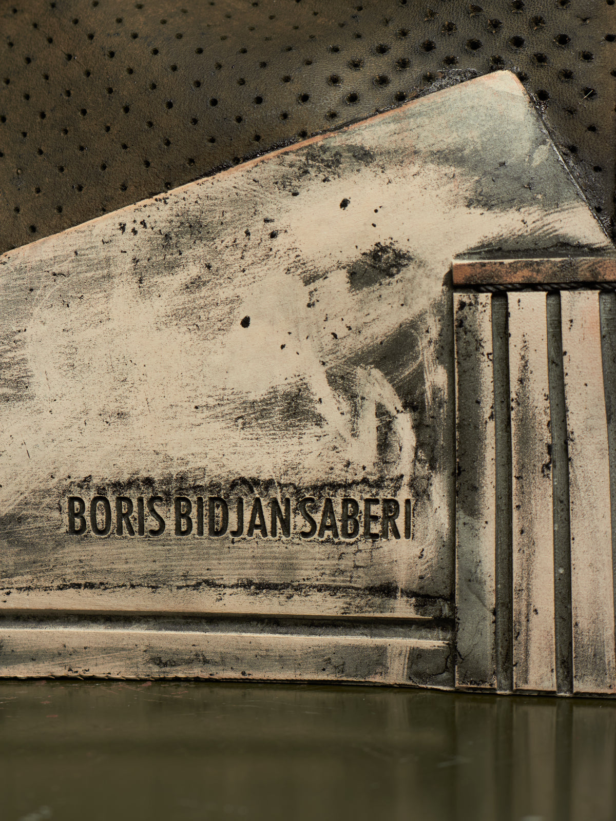 A close-up of a distressed, weathered metallic surface with the text 'BORIS BIDJAN SABERI' embossed or printed across the center, set against a dark perforated background.