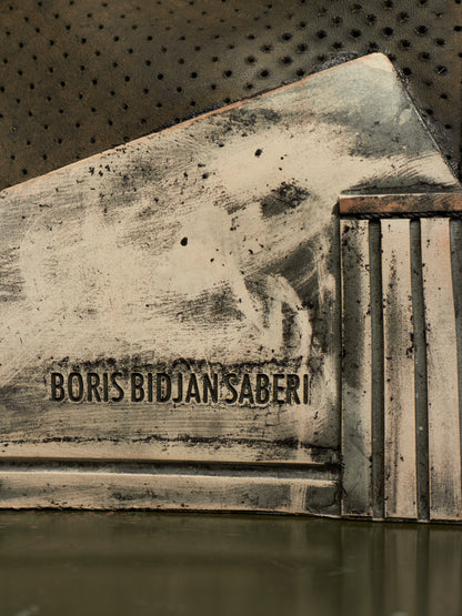 A close-up of a distressed, weathered metallic surface with the text 'BORIS BIDJAN SABERI' embossed or printed across the center, set against a dark perforated background.