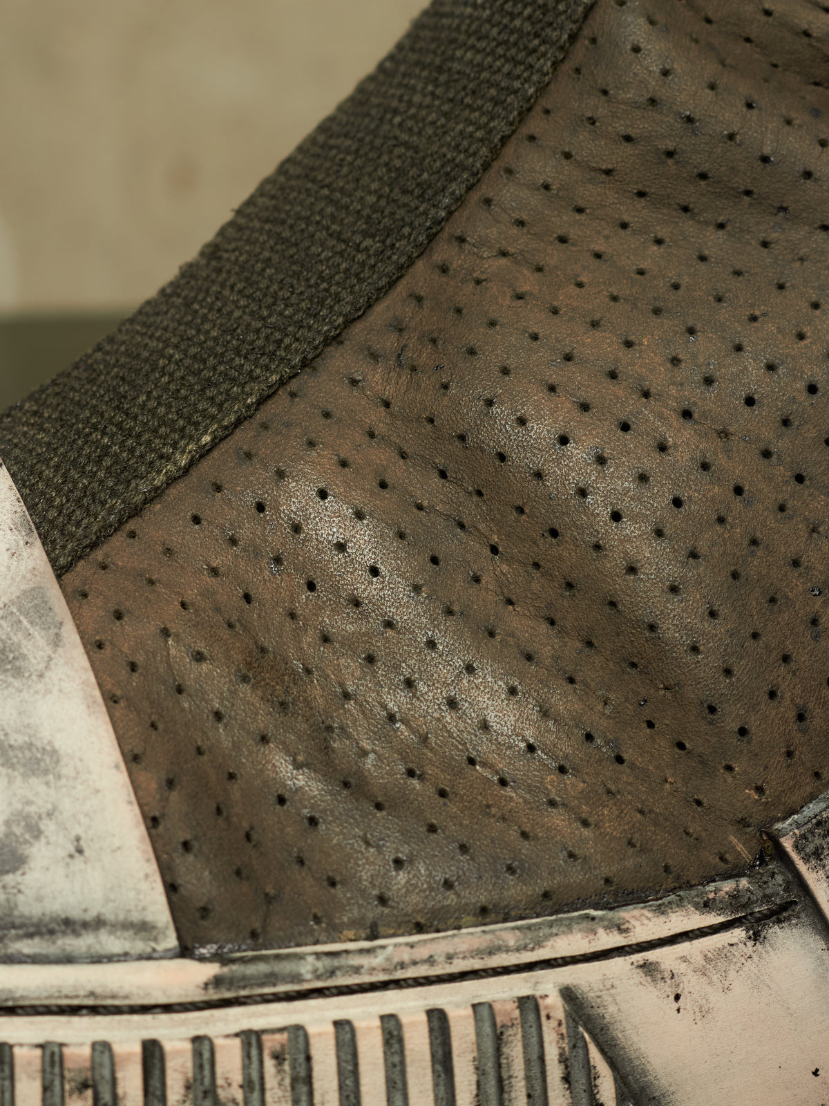 Close-up detail of a distressed leather shoe or boot upper, showing perforated brown leather, a dark olive fabric strap, and a heavily weathered off-white sole.