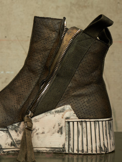 A close-up of a dark brown perforated leather ankle boot with a wide dark green elastic side strap and a chunky distressed white and gray platform sole.