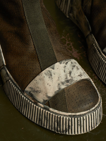 A close-up of a single worn open-toe slip-on shoe with a dark brown perforated upper, an olive green strap, and a heavily scuffed off-white and gray toe cap over a ribbed dark sole.