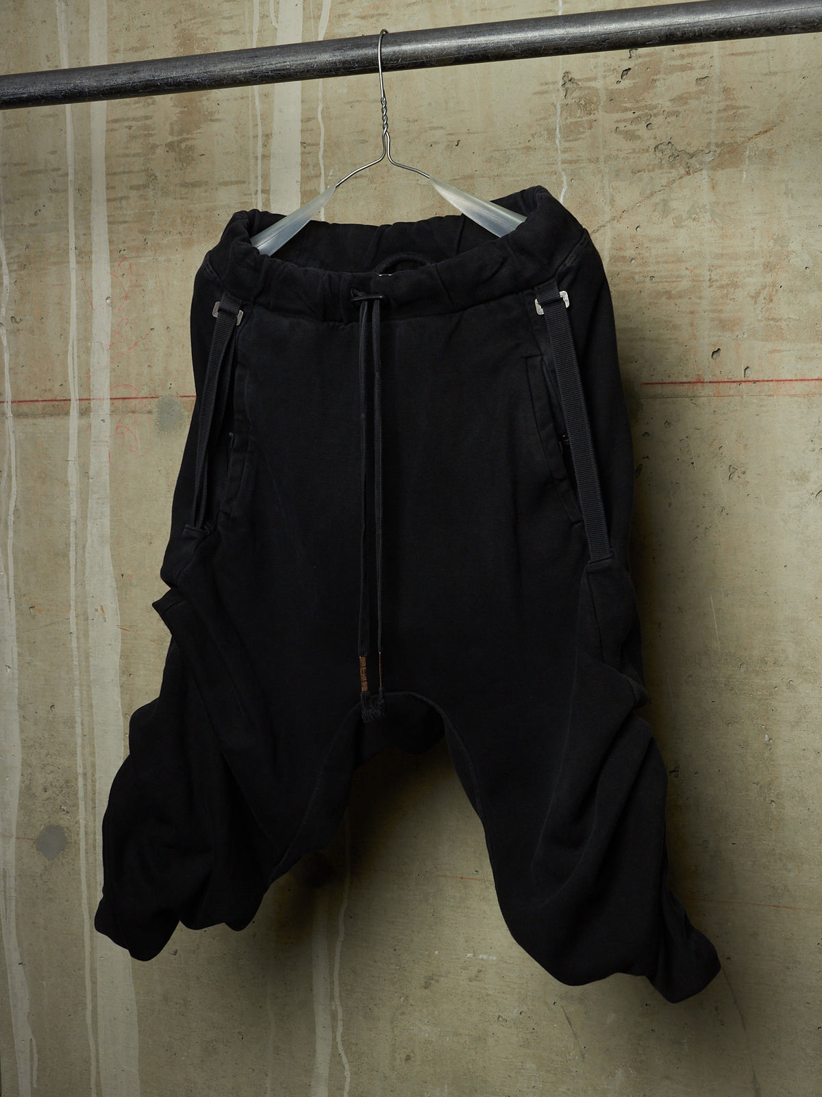 Black baggy sweatpants with a drawstring waist and side zipper details, hanging on a clear hanger from a metal rod against a textured concrete wall.