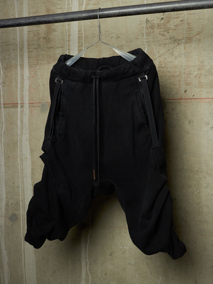 Black baggy sweatpants with a drawstring waist and side zipper details, hanging on a clear hanger from a metal rod against a textured concrete wall.