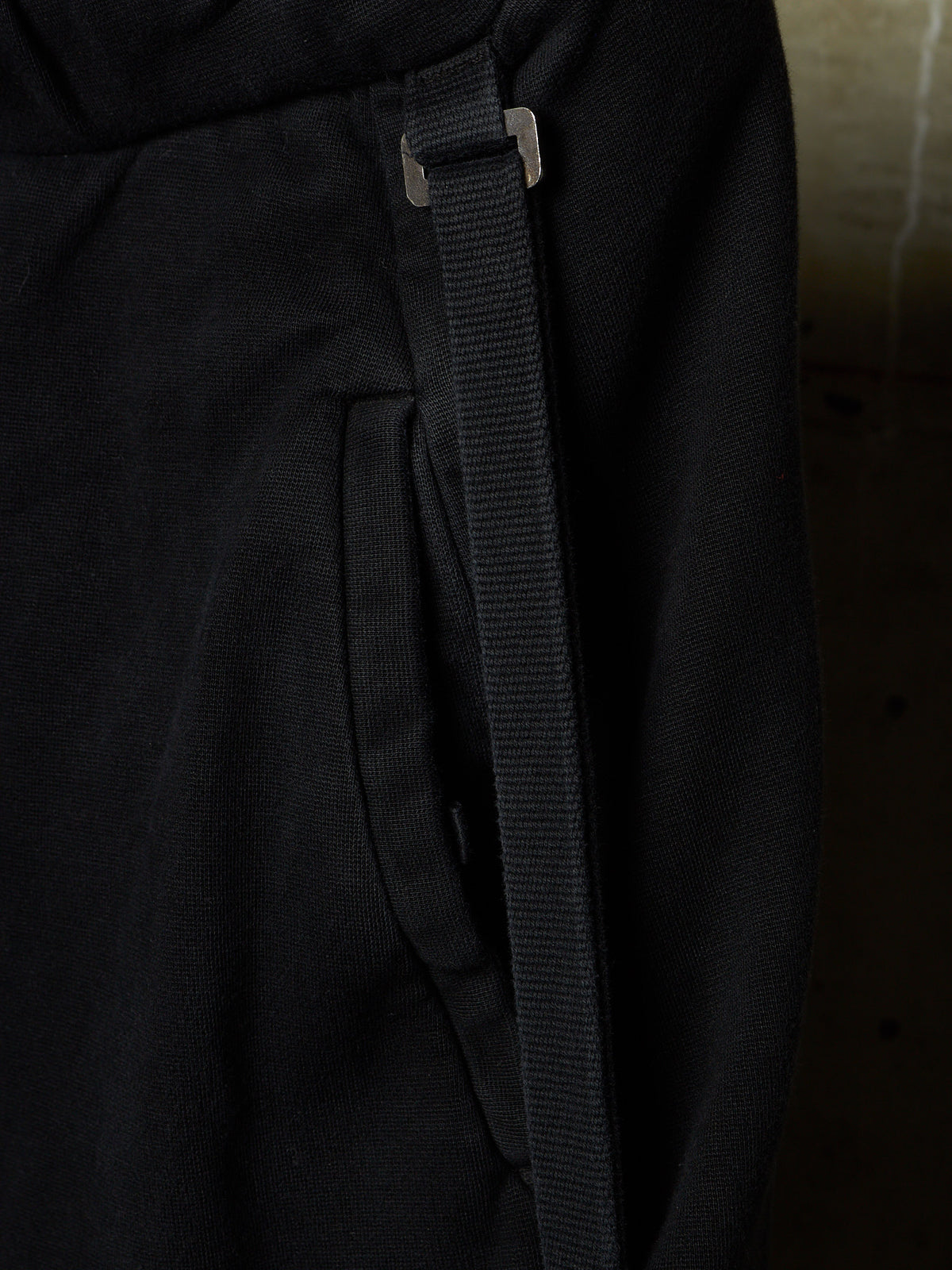 Close-up detail of black fabric with a vertical black strap and a small silver metal slider, shown against a dark background.