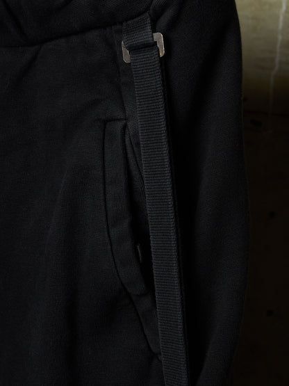 Close-up detail of black fabric with a vertical black strap and a small silver metal slider, shown against a dark background.