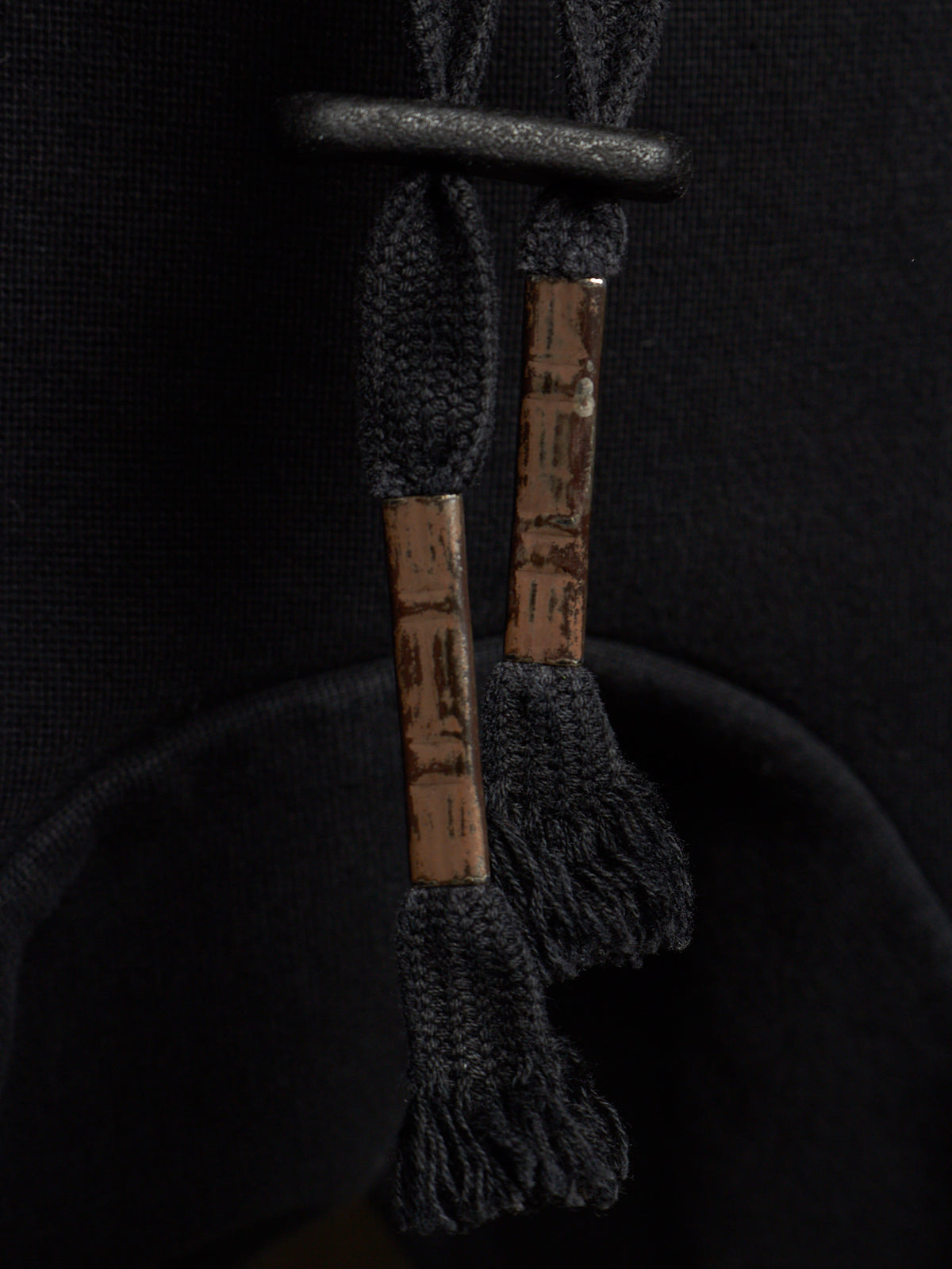 Close-up of a black drawstring bag with textured brown wooden aglets and black frayed tassel ends, set against a dark black fabric background.