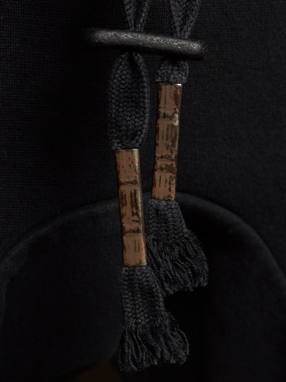 Close-up of a black drawstring bag with textured brown wooden aglets and black frayed tassel ends, set against a dark black fabric background.