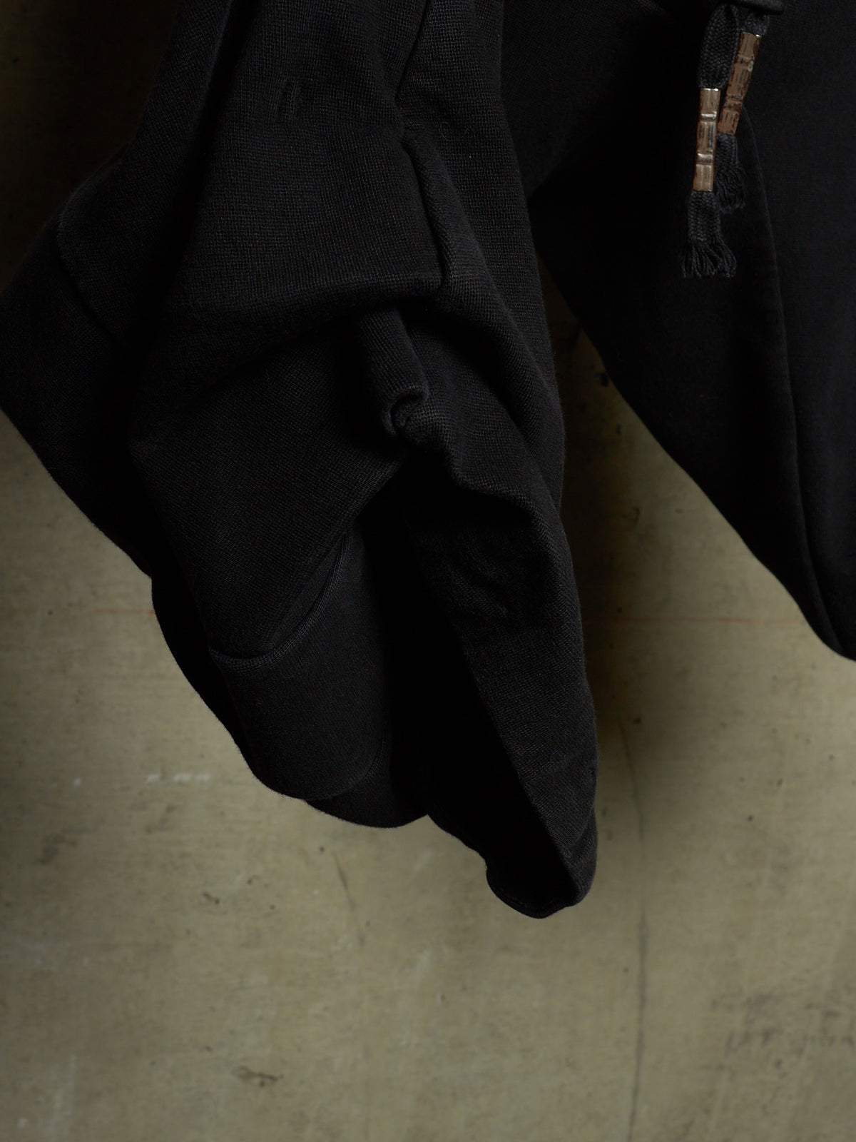 A close-up of a black garment, likely a jacket or coat, showing a textured fabric surface and a zipper detail, set against a concrete floor.