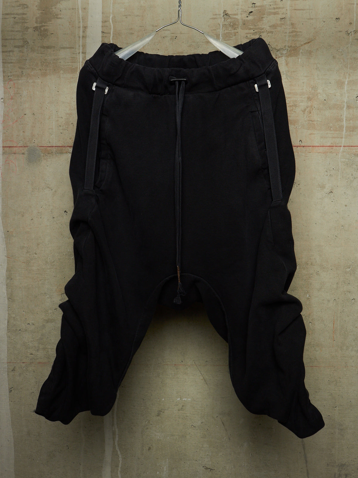 Black harem-style pants with a drawstring waist and side zipper details, hanging on a wire hanger against a textured concrete wall.
