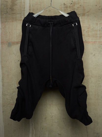 Black harem-style pants with a drawstring waist and side zipper details, hanging on a wire hanger against a textured concrete wall.