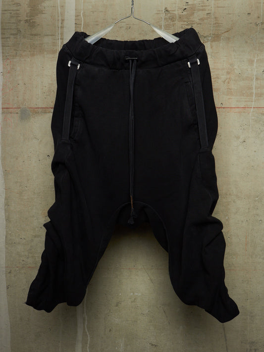 Black harem-style pants with a drawstring waist and side zipper details, hanging on a wire hanger against a textured concrete wall.