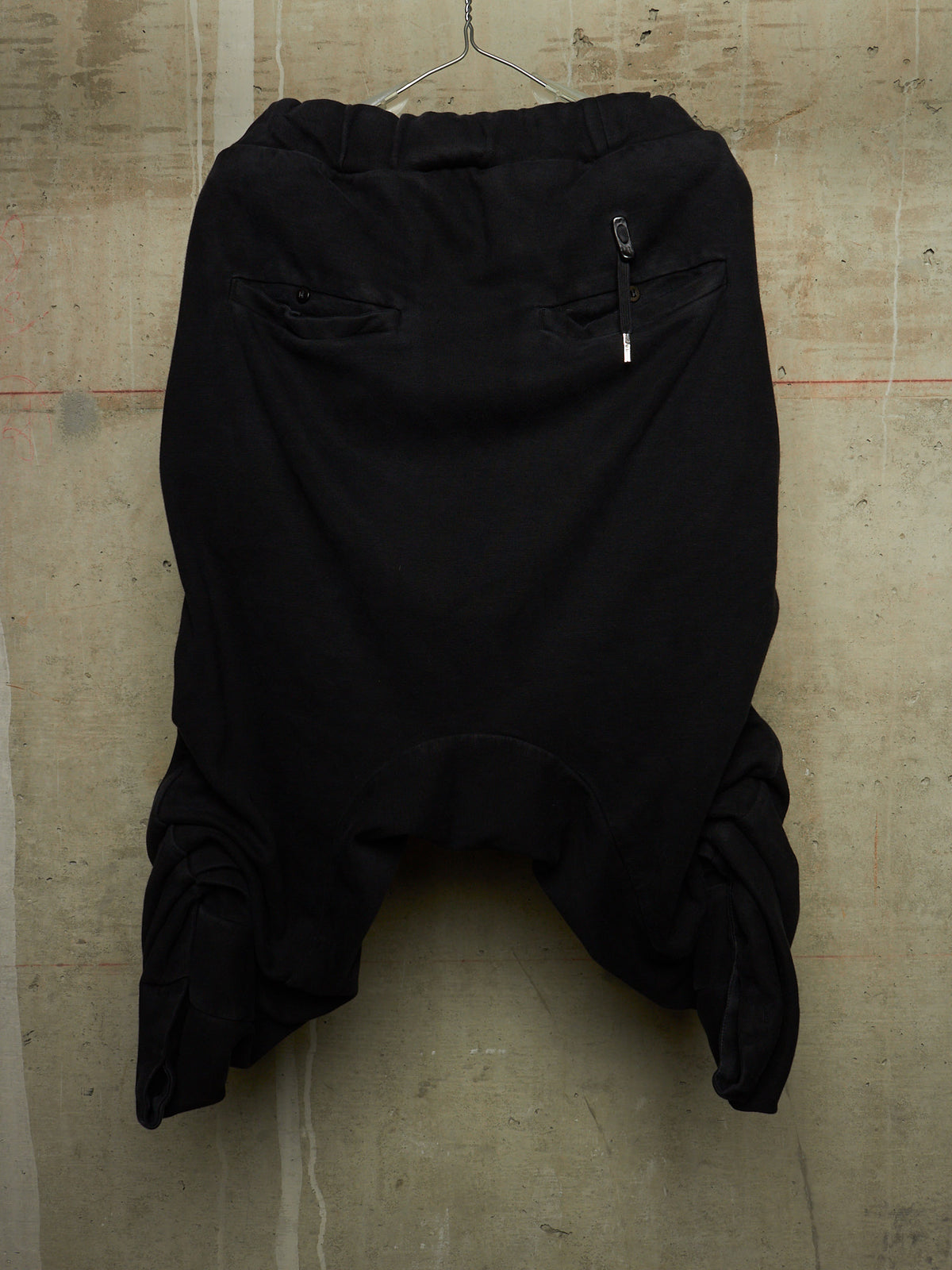 Black harem-style pants hanging against a textured concrete wall, featuring a gathered elastic waistband, side zip pocket, and voluminous draped legs.