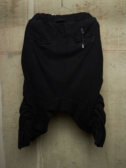 Black harem-style pants hanging against a textured concrete wall, featuring a gathered elastic waistband, side zip pocket, and voluminous draped legs.