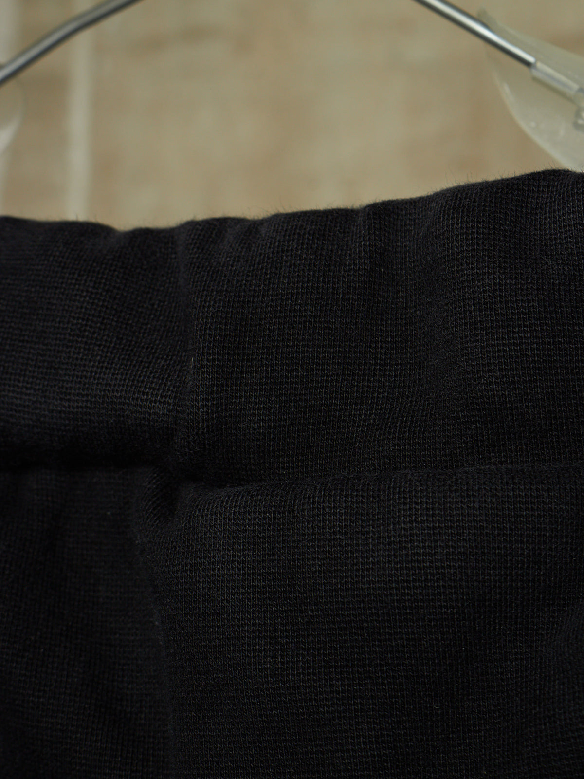 Close-up of a black textured fabric garment, likely a sweater or knit top, hanging on a clear plastic hanger against a beige background.