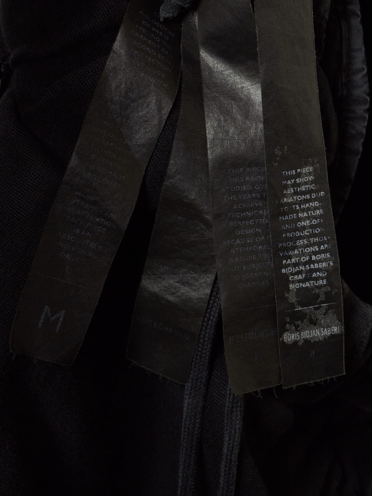 Close-up of a black garment with three wide black fabric straps or tags hanging down, printed with white text and a small white logo near the bottom right.