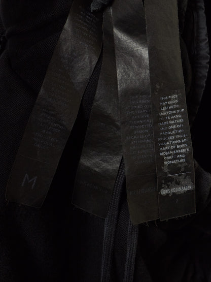 Close-up of a black garment with three wide black fabric straps or tags hanging down, printed with white text and a small white logo near the bottom right.
