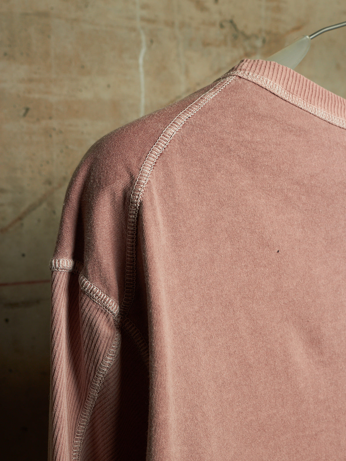 A close-up of a dusty rose pink long-sleeve sweatshirt with ribbed raglan seams and a ribbed crew neck, hanging on a clear plastic hanger against a textured beige wall.