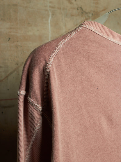 A close-up of a dusty rose pink long-sleeve sweatshirt with ribbed raglan seams and a ribbed crew neck, hanging on a clear plastic hanger against a textured beige wall.