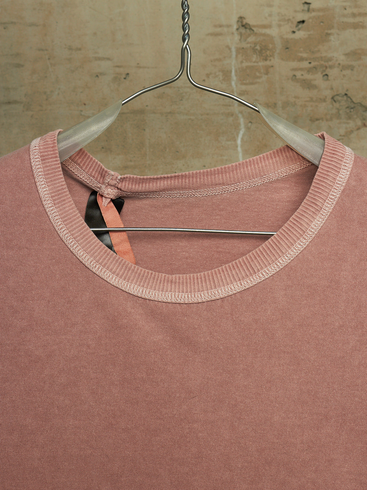 A dusty rose pink knit top with a contrasting light beige ribbed crew neck, hanging on a clear plastic hanger against a textured beige brick wall.