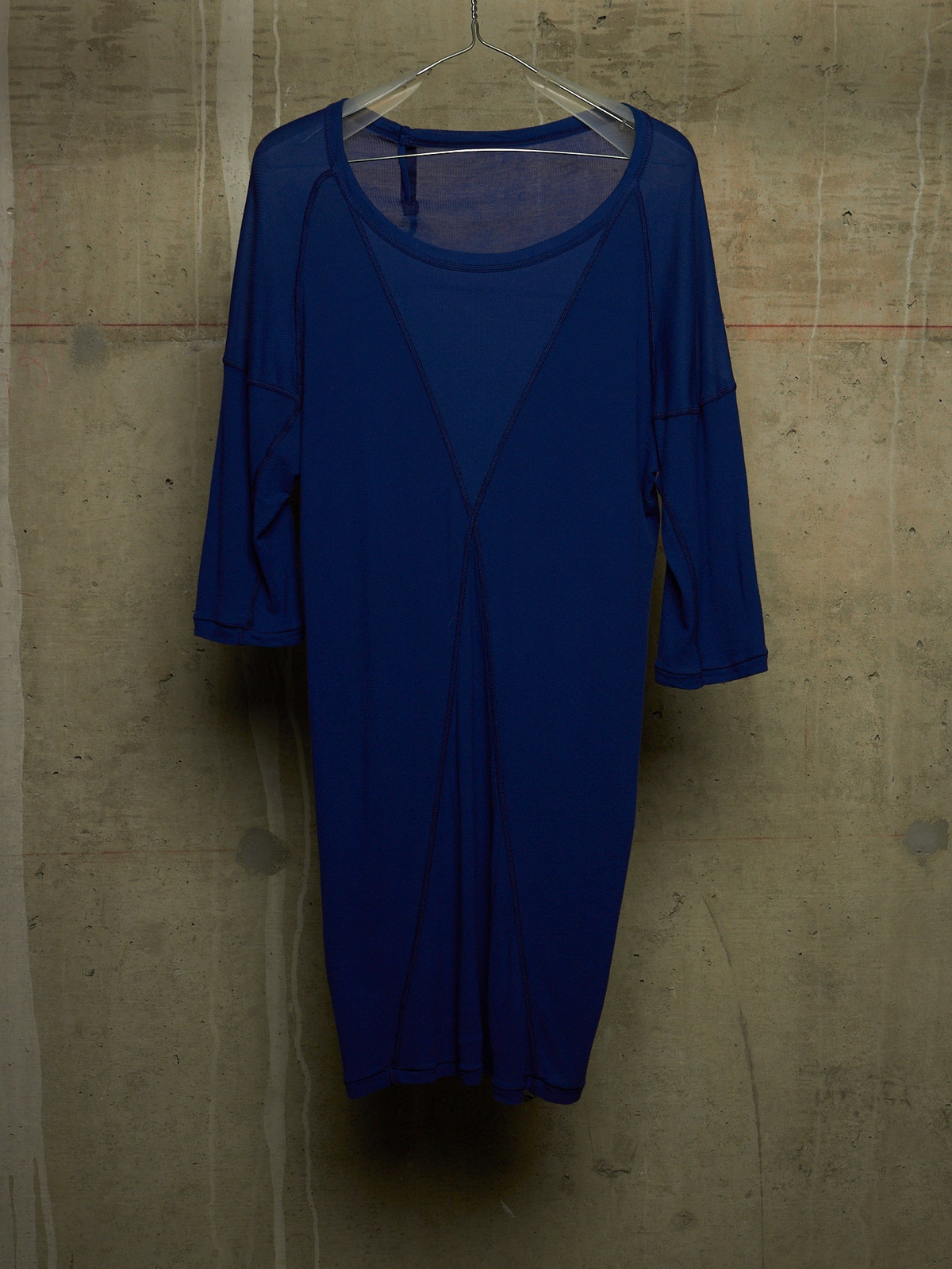A royal blue sheer mesh dress with three-quarter sleeves and a scoop neckline, hanging on a clear hanger against a textured concrete wall.