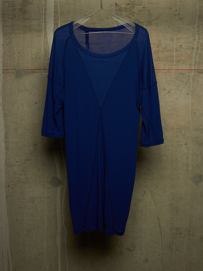 A royal blue sheer mesh dress with three-quarter sleeves and a scoop neckline, hanging on a clear hanger against a textured concrete wall.