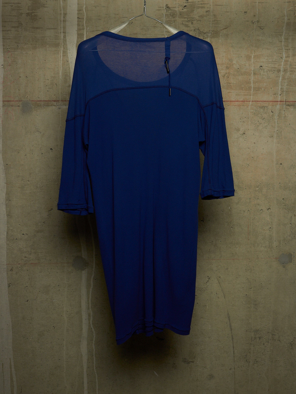 A royal blue sheer mesh long-sleeve dress with a solid blue front panel, hanging on a metal hanger against a textured concrete wall.