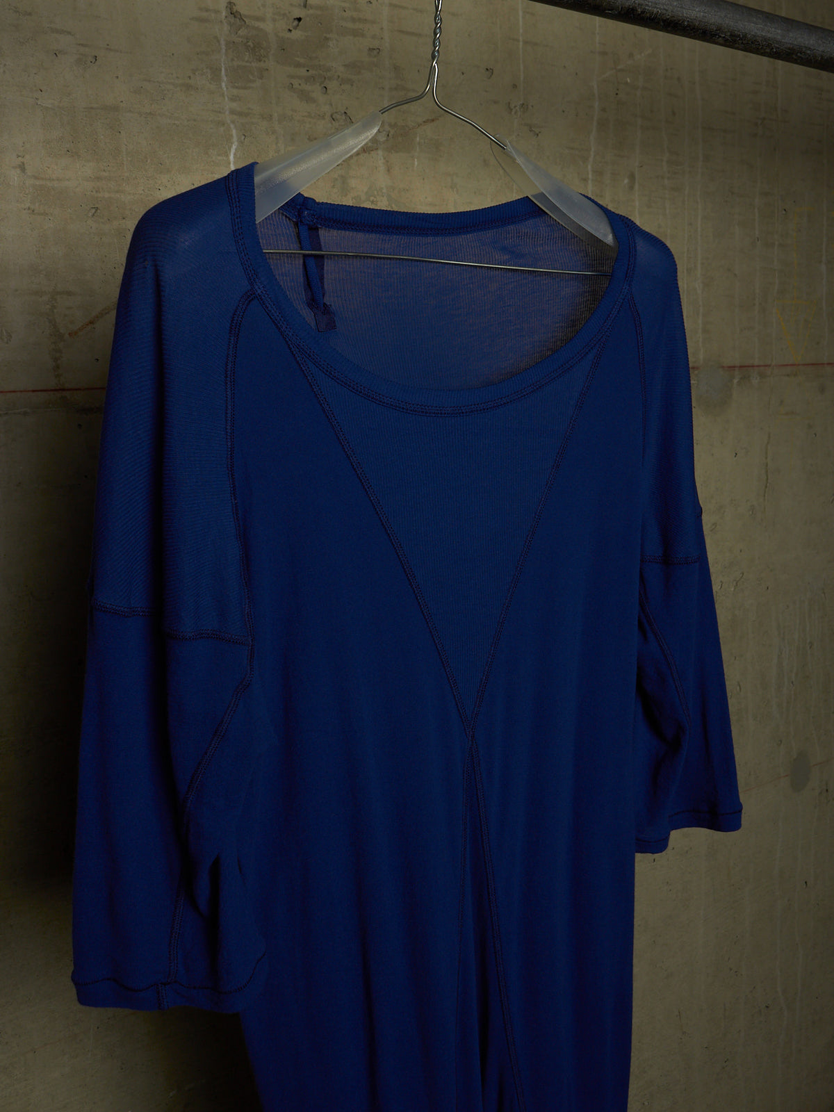 A royal blue long-sleeve top with a scoop neckline, hanging on a clear plastic hanger against a textured concrete wall.