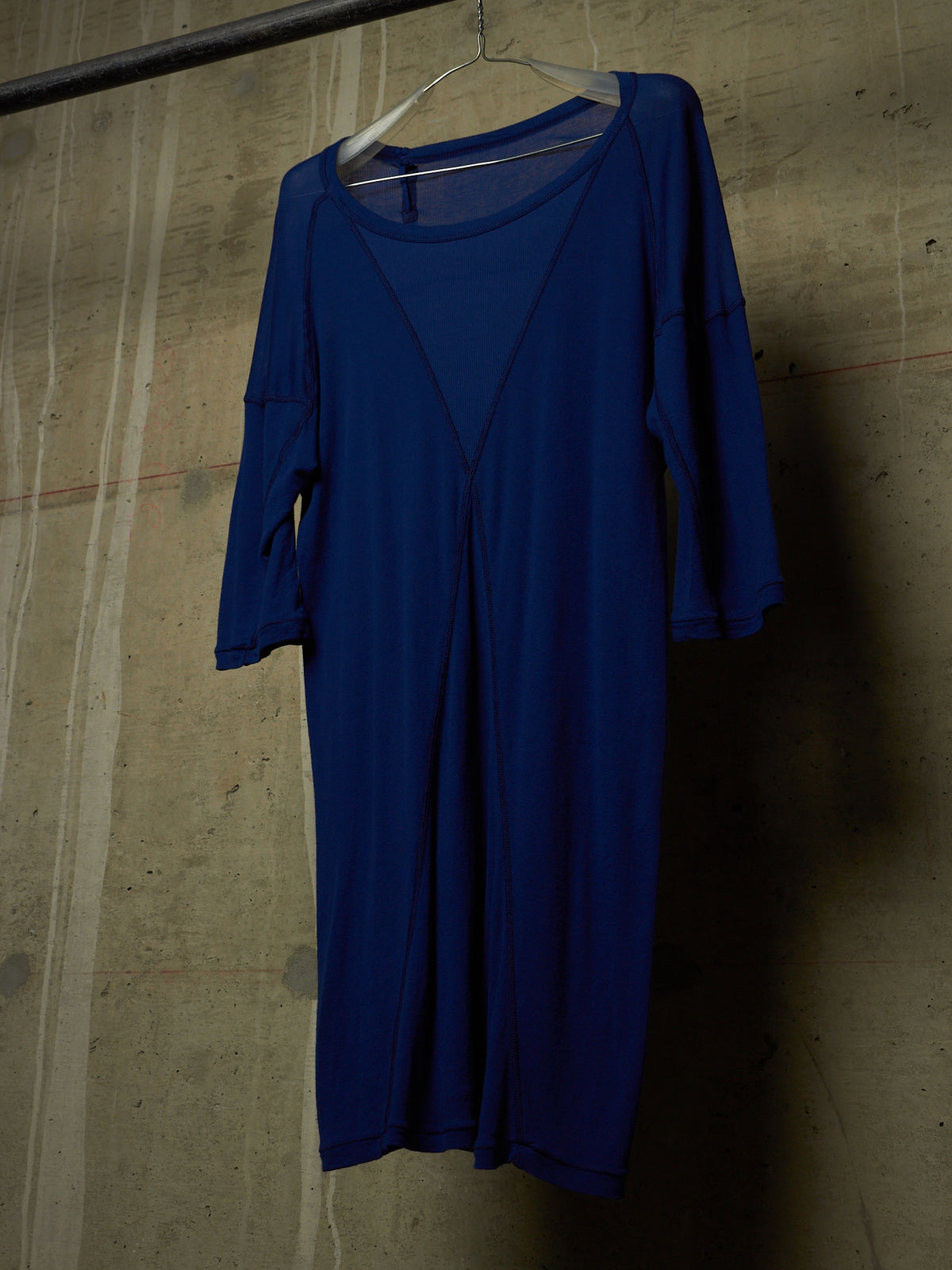 A royal blue long-sleeve dress hanging on a clear plastic hanger against a textured concrete wall.