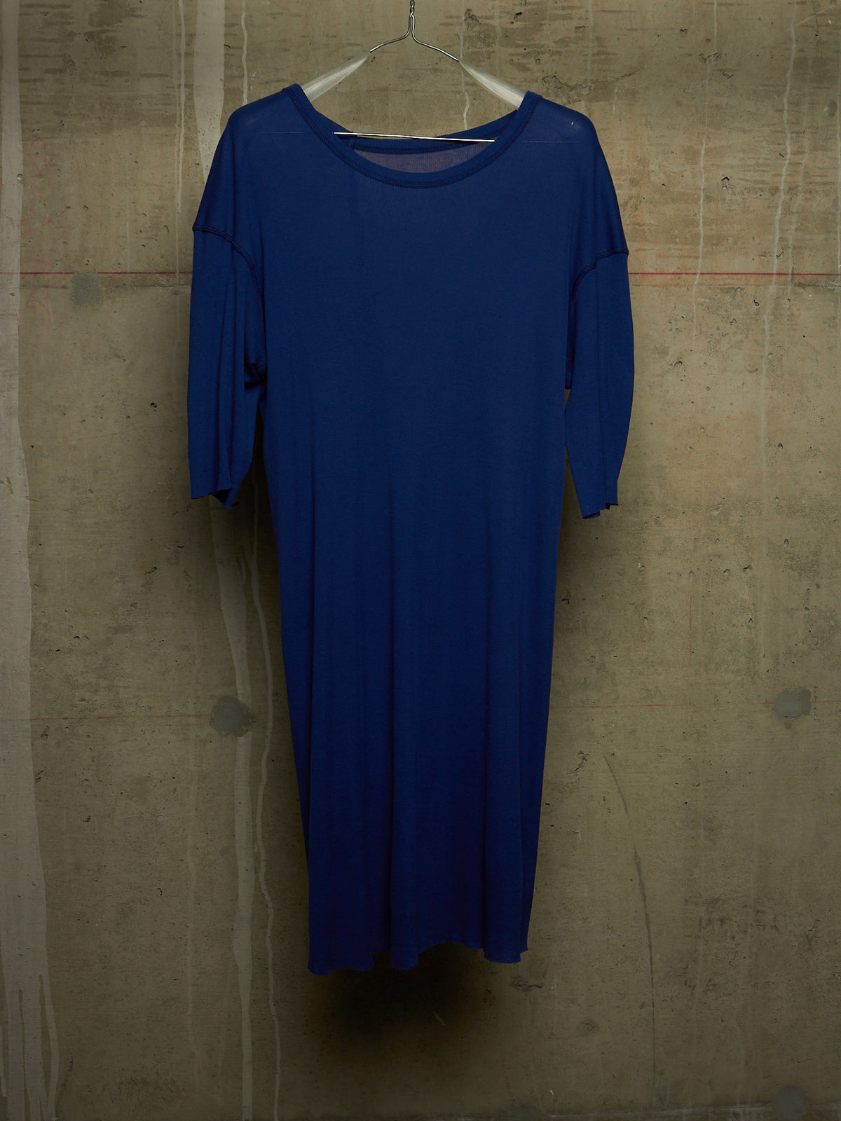 A royal blue long-sleeve dress hanging on a clear plastic hanger against a textured concrete wall.