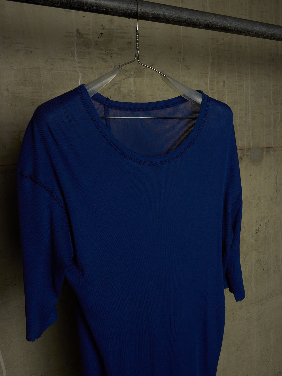 A royal blue short-sleeve top hanging on a clear plastic hanger against a textured concrete wall.