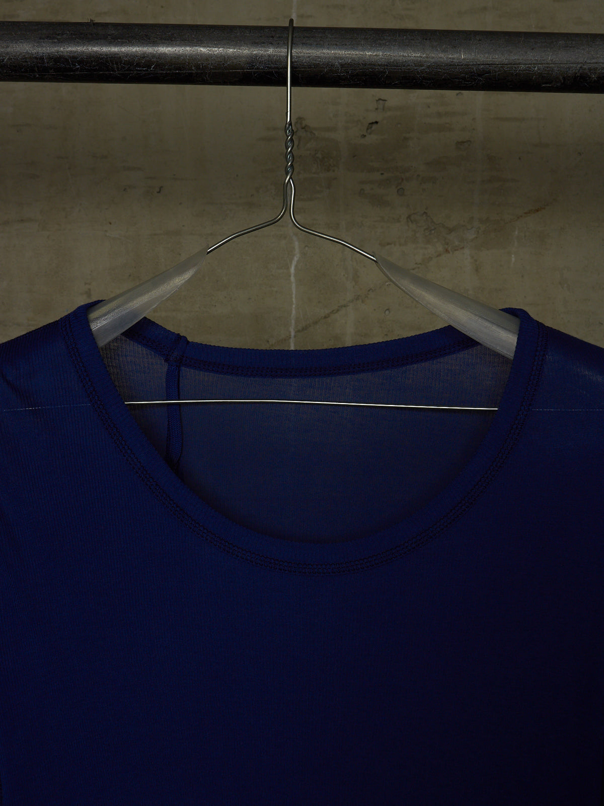 A close-up of a dark blue scoop-neck top hanging on a metal hanger from a dark horizontal bar against a textured concrete wall.