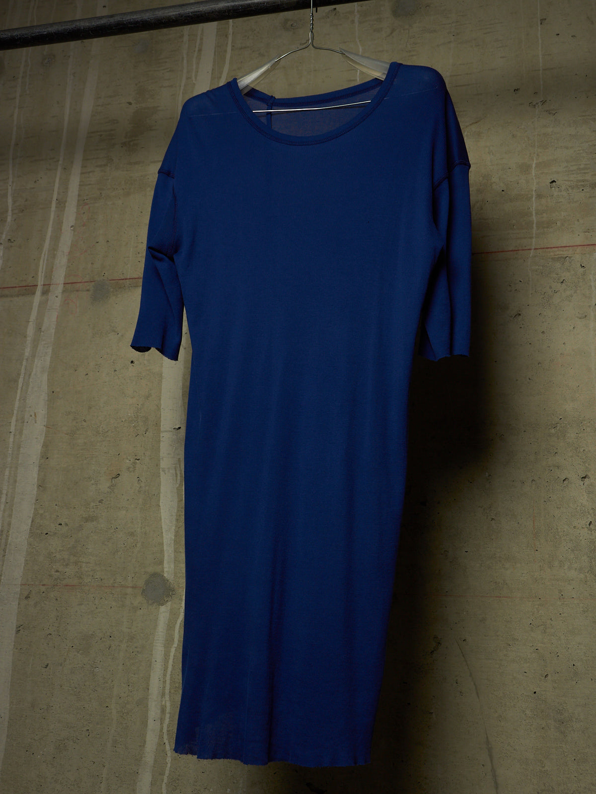 A royal blue short-sleeve dress hanging on a white plastic hanger against a textured concrete wall.