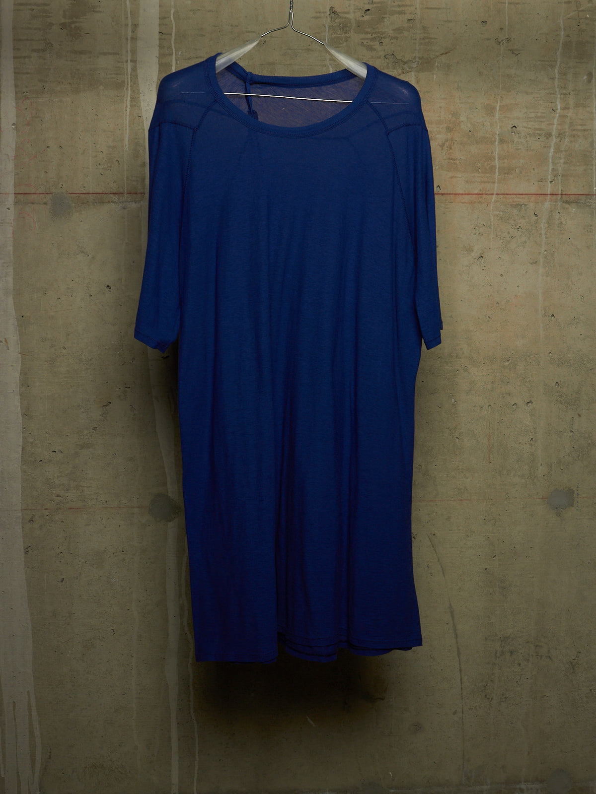 A royal blue short-sleeve dress with sheer shoulder panels, hanging on a white hanger against a textured concrete wall.