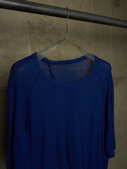 A royal blue long-sleeve knit top hanging on a clear plastic hanger from a dark metal rod against a textured concrete wall.