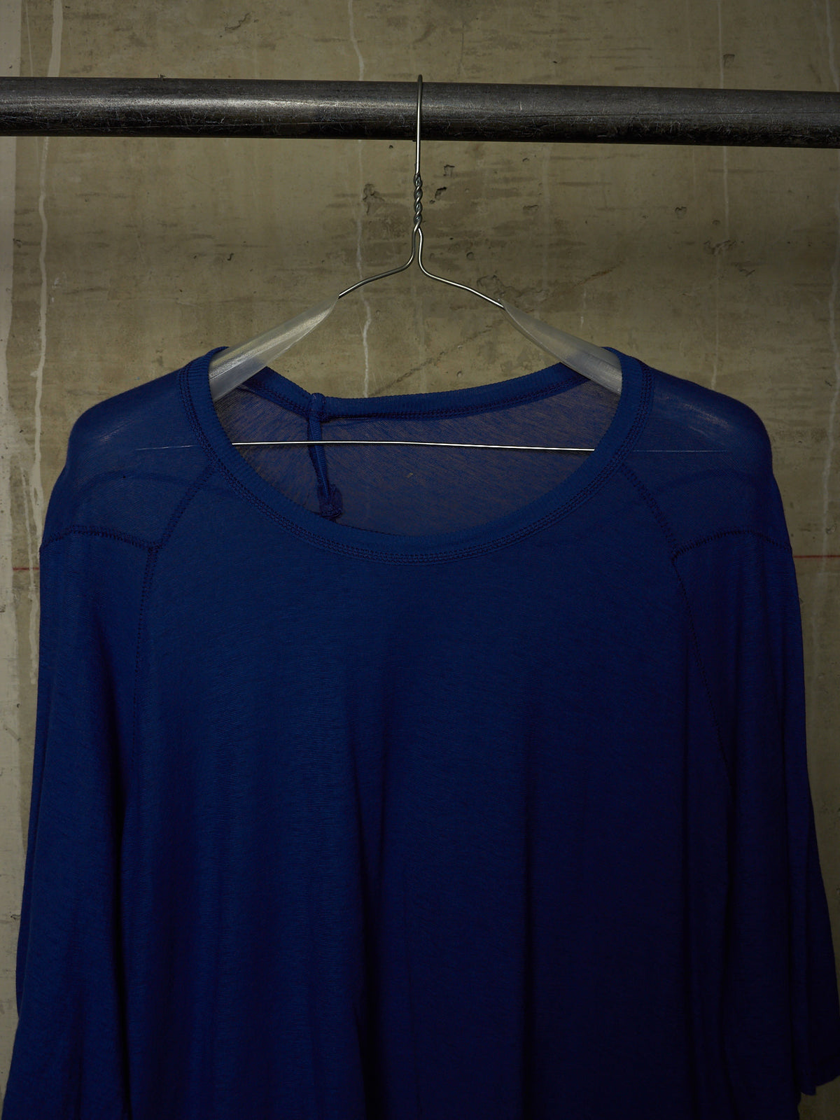 A royal blue long-sleeve top with a sheer mesh shoulder overlay, hanging on a clear plastic hanger from a dark metal clothing rack against a textured concrete wall.