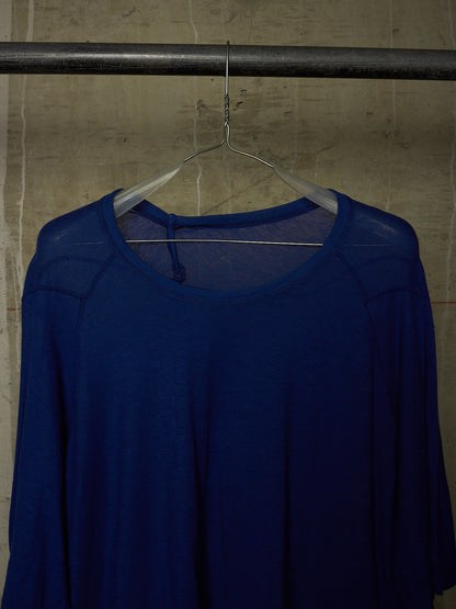 A royal blue long-sleeve top with a sheer mesh shoulder overlay, hanging on a clear plastic hanger from a dark metal clothing rack against a textured concrete wall.