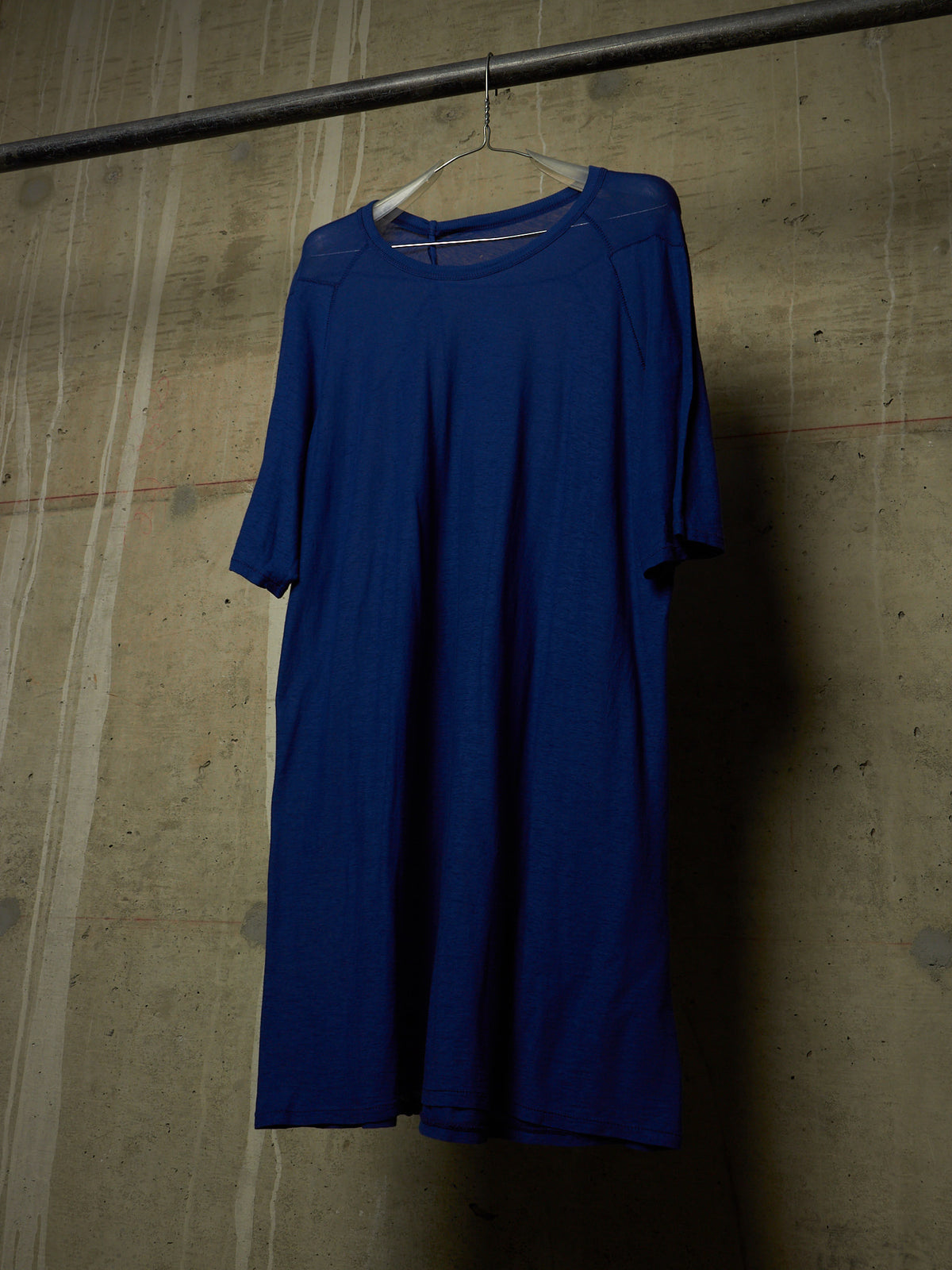 A royal blue short-sleeve dress hanging on a white hanger from a dark metal rod against a textured concrete wall.