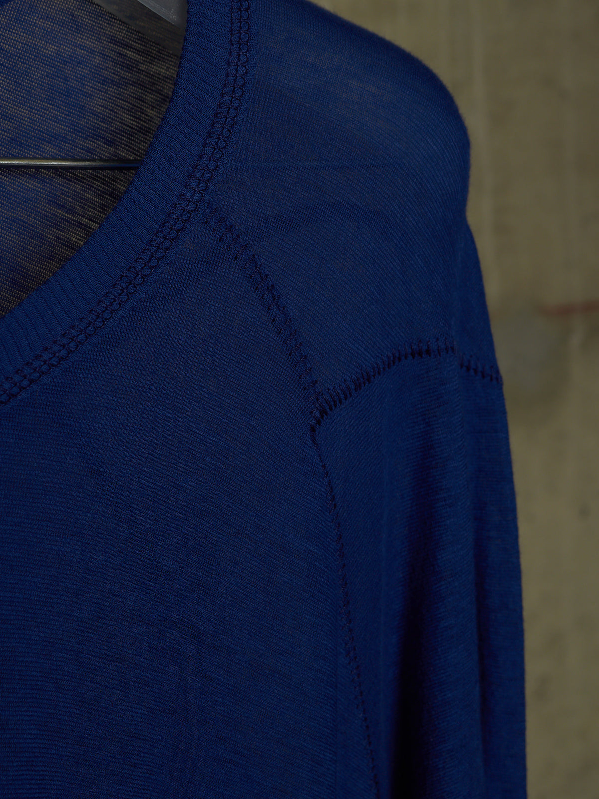 Close-up of a royal blue knit sweater on a hanger, showing the ribbed v-neck collar and shoulder seam details against a neutral textured background.